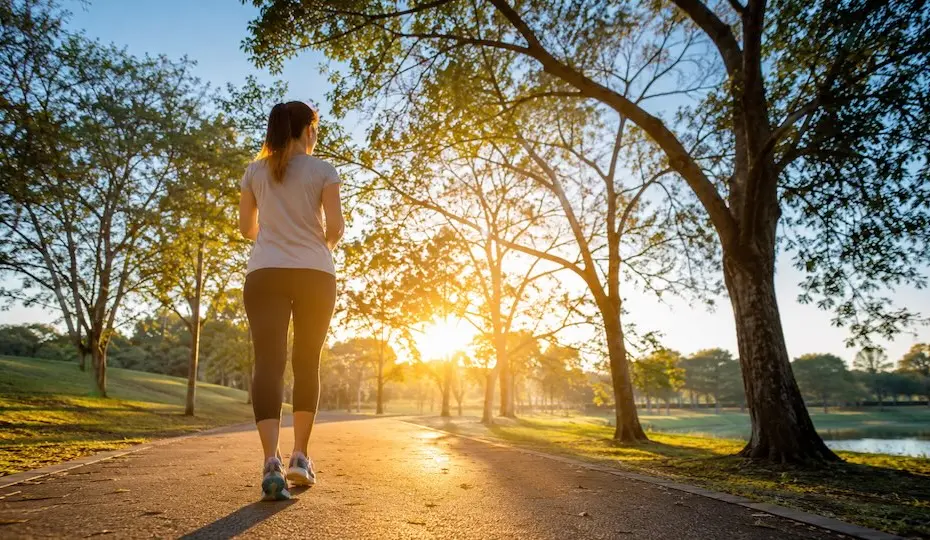 Pessoa caminhando em um parque arborizado durante o nascer do sol, representando hábitos saudáveis e qualidade de vida
