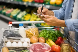 Pessoa faz lista de compras diante de carrinho com feijão, leite, carne e outros alimentos no supermercado