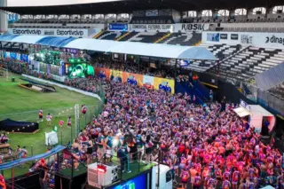 Multidão de foliões reunida na pista de um estádio em frente a trios elétricos, com arquibancadas ao fundo