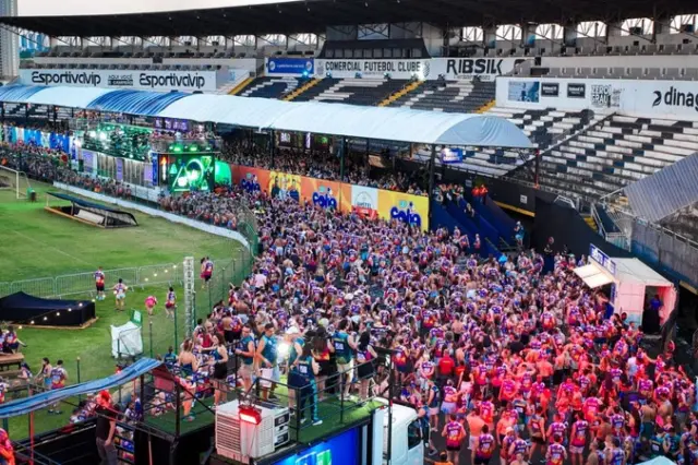 Multidão de foliões reunida na pista de um estádio em frente a trios elétricos, com arquibancadas ao fundo