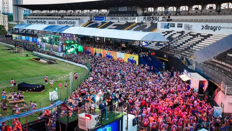 Multidão de foliões reunida na pista de um estádio em frente a trios elétricos, com arquibancadas ao fundo