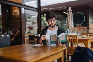 Homem com barba e óculos sentado em mesa de cafeteria ao ar livre, segurando uma xícara e lendo um livro