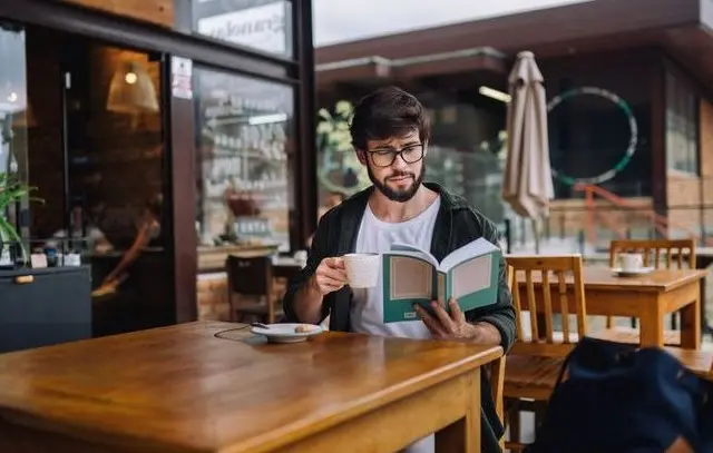 Homem com barba e óculos sentado em mesa de cafeteria ao ar livre, segurando uma xícara e lendo um livro