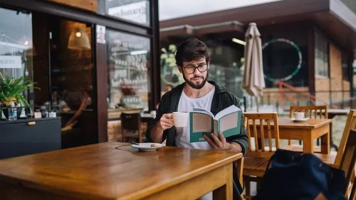 Homem com barba e óculos sentado em mesa de cafeteria ao ar livre, segurando uma xícara e lendo um livro