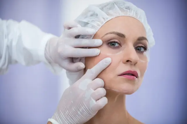 mulher de meia-idade com touca cirúrgica e marcações brancas sob os olhos enquanto mãos enluvadas tocam sua face