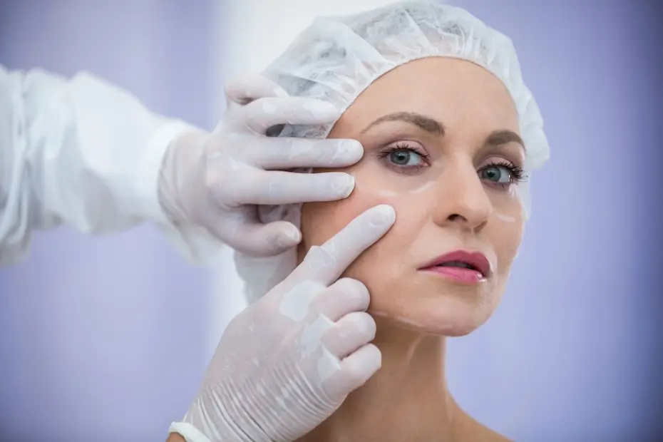 mulher de meia-idade com touca cirúrgica e marcações brancas sob os olhos enquanto mãos enluvadas tocam sua face