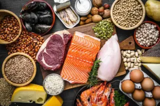 Alimentos ricos em proteínas e minerais dispostos sobre superfície escura, incluindo salmão, carne vermelha, peito de frango, ovos, queijo, camarões, mariscos, leguminosas, nozes e sementes