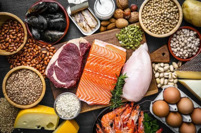 Alimentos ricos em proteínas e minerais dispostos sobre superfície escura, incluindo salmão, carne vermelha, peito de frango, ovos, queijo, camarões, mariscos, leguminosas, nozes e sementes