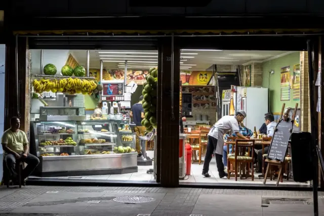 fachada aberta de um pequeno estabelecimento comercial à noite com balcão de frutas, bananas penduradas, uma pessoa sentada na calçada e funcionários atendendo clientes