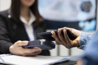 Pessoa realizando pagamento com celular sobre maquininha de cartão; mão segura smartphone próximo ao POS e outra segura o aparelho, com pessoa ao fundo desfocada