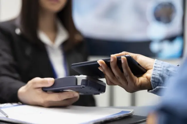 Pessoa realizando pagamento com celular sobre maquininha de cartão; mão segura smartphone próximo ao POS e outra segura o aparelho, com pessoa ao fundo desfocada