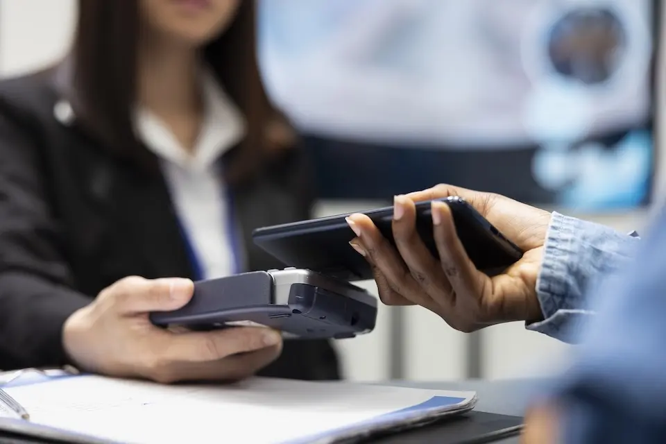 Pessoa realizando pagamento com celular sobre maquininha de cartão; mão segura smartphone próximo ao POS e outra segura o aparelho, com pessoa ao fundo desfocada