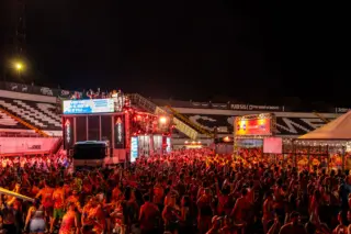 Multidão de foliões à noite diante de um trio elétrico/truck de show iluminado, com arquibancadas ao fundo