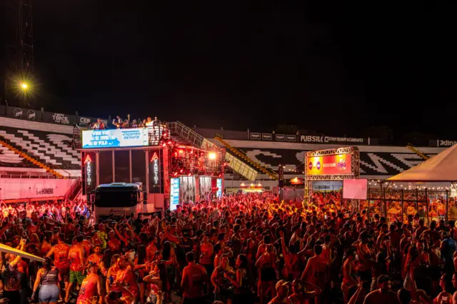 Multidão de foliões à noite diante de um trio elétrico/truck de show iluminado, com arquibancadas ao fundo