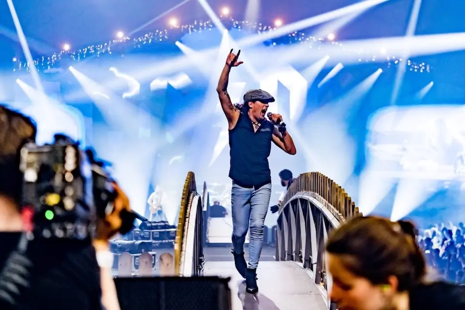 vocalista caminhando por uma passarela de palco, com microfone na mão e luzes de cena ao fundo