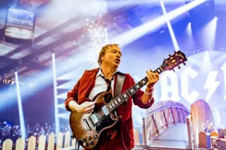 músico com blazer vermelho tocando guitarra elétrica em primeiro plano, com luzes de palco e parte do letreiro AC/DC ao fundo
