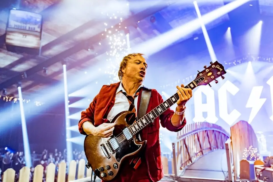 músico com blazer vermelho tocando guitarra elétrica em primeiro plano, com luzes de palco e parte do letreiro AC/DC ao fundo