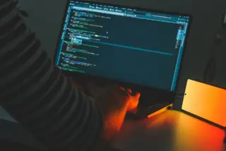 mãos digitando em notebook com editor de código na tela e smartphone com luz laranja ao lado