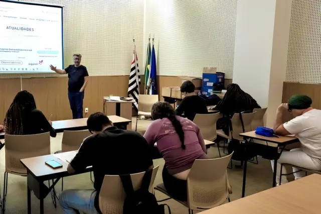 Sala de aula com adultos sentados em carteiras enquanto um instrutor em pé aponta para uma tela projetada ao fundo