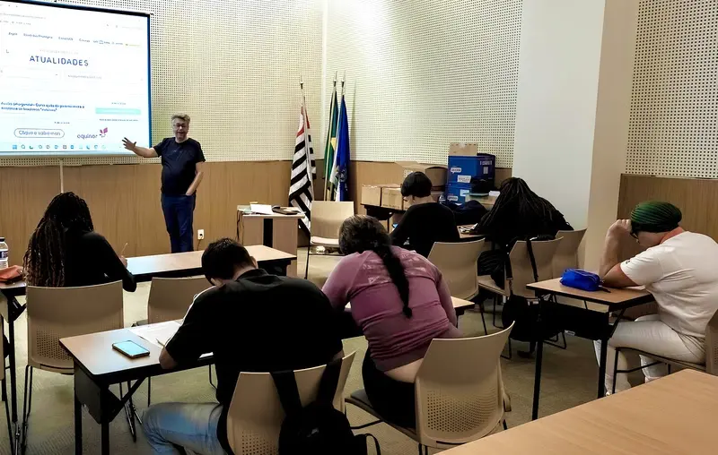 Sala de aula com adultos sentados em carteiras enquanto um instrutor em pé aponta para uma tela projetada ao fundo