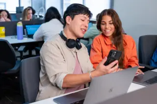 Dois jovens sentados lado a lado em mesas com laptops; um rapaz segura um celular e mostra a tela para uma jovem que sorri