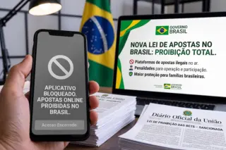 Mão segurando celular com tela que indica 'APLICATIVO BLOQUEADO. APOSTAS ONLINE PROIBIDAS NO BRASIL. Acesso Encerrado.' Monitor ao fundo mostra 'NOVA LEI DE APOSTAS NO BRASIL: PROIBIÇÃO TOTAL.' Jornal 'Diário Oficial da União' sobre a mesa; bandeira do Brasil ao fundo.