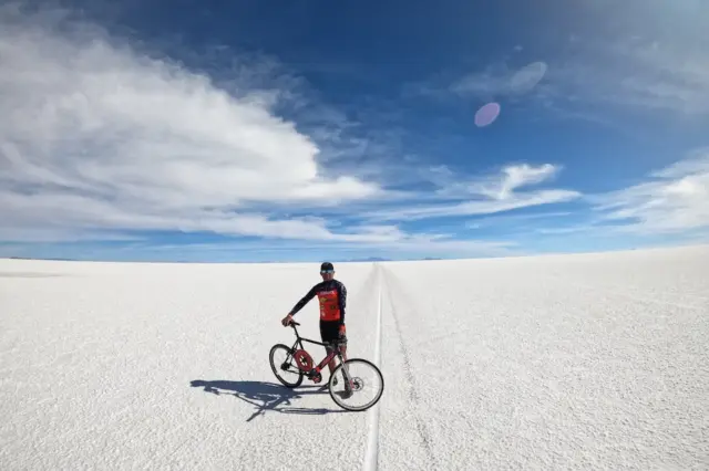 Ciclista parado ao lado de bicicleta sobre o Salar de Uyuni com céu azul e nuvens
