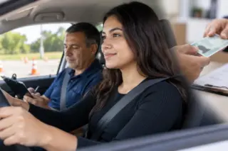 Mulher jovem sentada ao volante usando cinto de segurança, com instrutor ao lado e uma mão segurando um cartão com foto