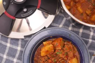 Tampa da Panela de Pressão Royal Prestige ao lado de panela aberta e tigela azul com carne de panela e batatas sobre pano xadrez