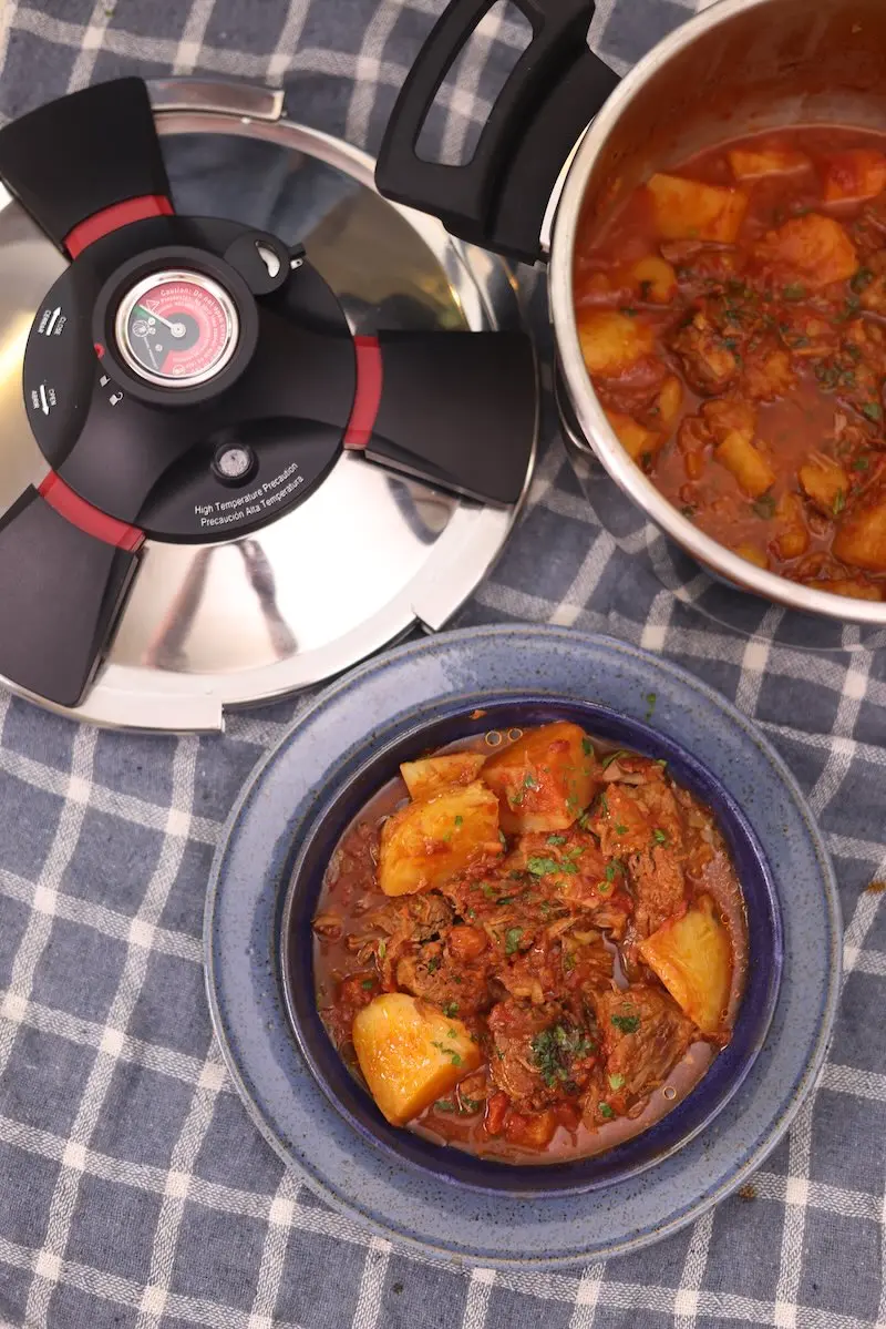 Tampa da Panela de Pressão Royal Prestige ao lado de panela aberta e tigela azul com carne de panela e batatas sobre pano xadrez