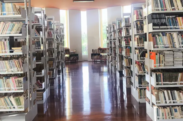 Corredor interno de biblioteca com estantes metálicas alinhadas e prateleiras cheias de livros