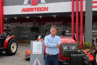 homem sorrindo com braços cruzados em frente a trator vermelho e estande com logotipo Agritech