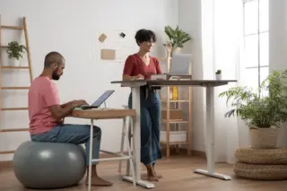 Homem sentado em bola de exercícios usando laptop e mulher em pé em mesa ajustável trabalhando em um espaço com plantas e estante