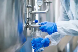 Mãos com luvas azuis abrindo válvula de tanque inox enquanto líquido é coletado em um béquer