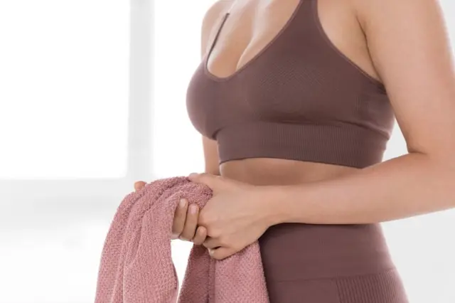 Tronco de mulher com top esportivo marrom e cintura alta visível, segurando uma toalha rosa com as duas mãos