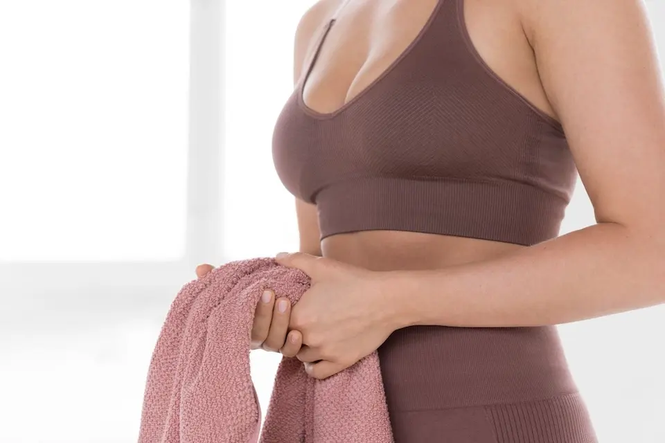 Tronco de mulher com top esportivo marrom e cintura alta visível, segurando uma toalha rosa com as duas mãos