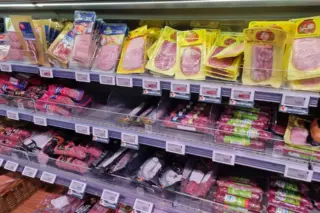 prateleira refrigerada com embalagens de embutidos e produtos cárneos processados fatiados e em pacotes, com etiquetas de preço visíveis