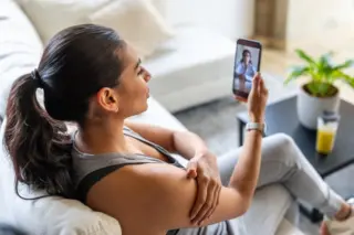 Mulher sentada de perfil em um sofá segurando um celular com uma vídeo-chamada de médico