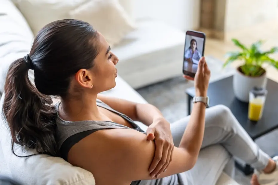 Mulher sentada de perfil em um sofá segurando um celular com uma vídeo-chamada de médico