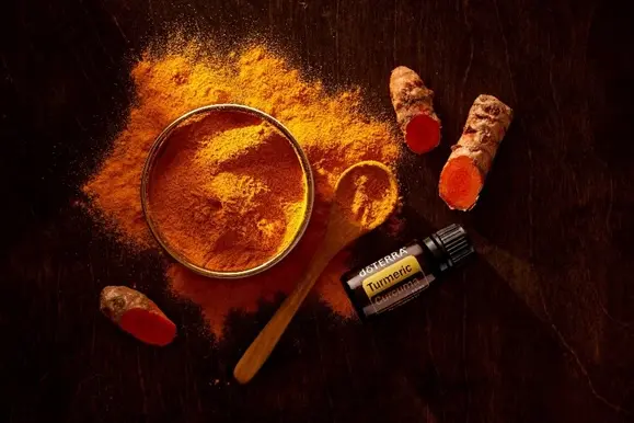 Tigela com pó de cúrcuma, colher de madeira com pó, raízes de cúrcuma cortadas e frasco pequeno rotulado 'Turmeric' sobre superfície escura