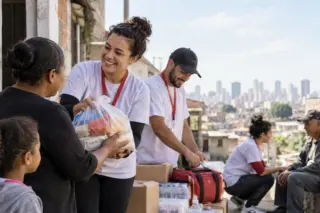 Voluntários de camisetas brancas entregam sacola com mantimentos a uma moradora, com criança observando; caixas e garrafas de água empilhadas e prédios da cidade ao fundo