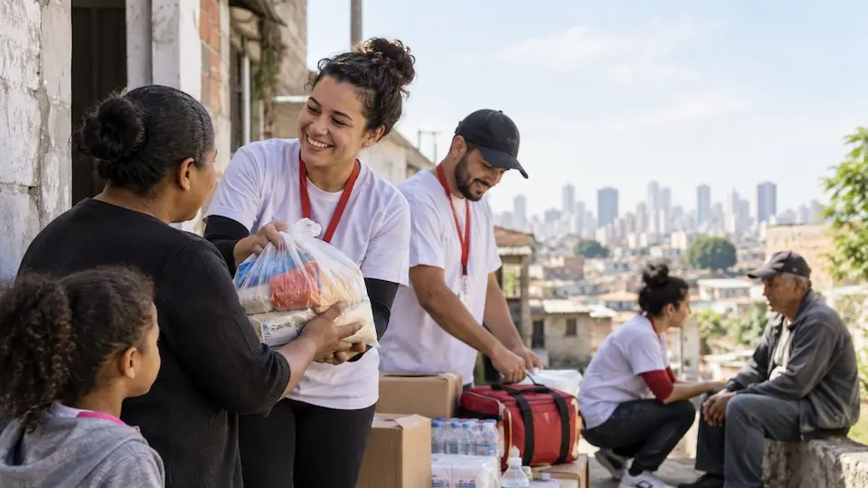 Voluntários de camisetas brancas entregam sacola com mantimentos a uma moradora, com criança observando; caixas e garrafas de água empilhadas e prédios da cidade ao fundo