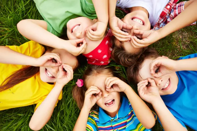 Cinco crianças deitadas sobre a grama formando um círculo, sorrindo e fazendo com as mãos uma forma de binóculo sobre os olhos