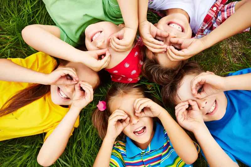 Cinco crianças deitadas sobre a grama formando um círculo, sorrindo e fazendo com as mãos uma forma de binóculo sobre os olhos