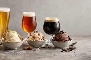Três taças de cerveja (clara, âmbar e escura) alinhadas atrás de três tigelas com bolas de sorvete, com pedaços de chocolate, nozes e uma fatia de limão em primeiro plano