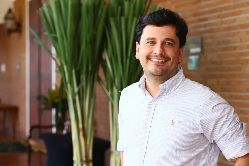 Homem adulto sorrindo com camisa social clara, em ambiente interno com plantas e parede de tijolos ao fundo