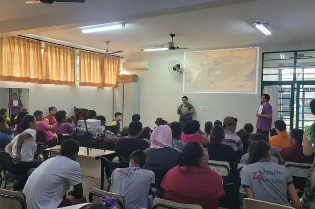Alunos e profissionais sentados em sala enquanto dois apresentadores fazem atividade com projeção na parede