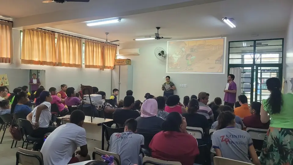 Alunos e profissionais sentados em sala enquanto dois apresentadores fazem atividade com projeção na parede