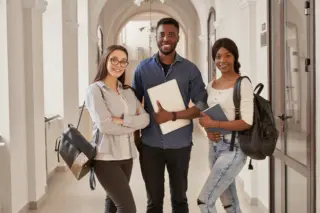 três estudantes com mochilas, um segurando um laptop e outro um caderno, em um corredor com arcadas