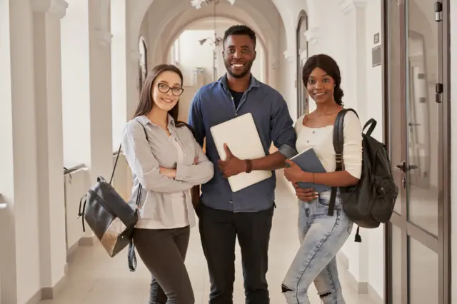 três estudantes com mochilas, um segurando um laptop e outro um caderno, em um corredor com arcadas